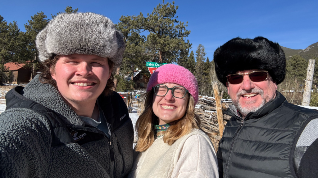 Three people smiling closely together outdoors on a sunny winter day, wearing warm jackets and fur hats. Snow-covered ground, pine trees, and mountains are visible in the background under a bright blue sky.