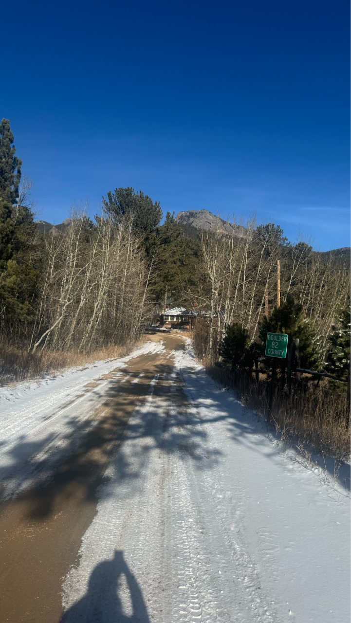 A snow-covered dirt road winding through a wooded mountain area with leafless trees and evergreens. A small cabin sits at the end of the road, mountains rise in the distance, and a sign on the right reads ‘Boulder County.’ The sky is clear and bright blue.